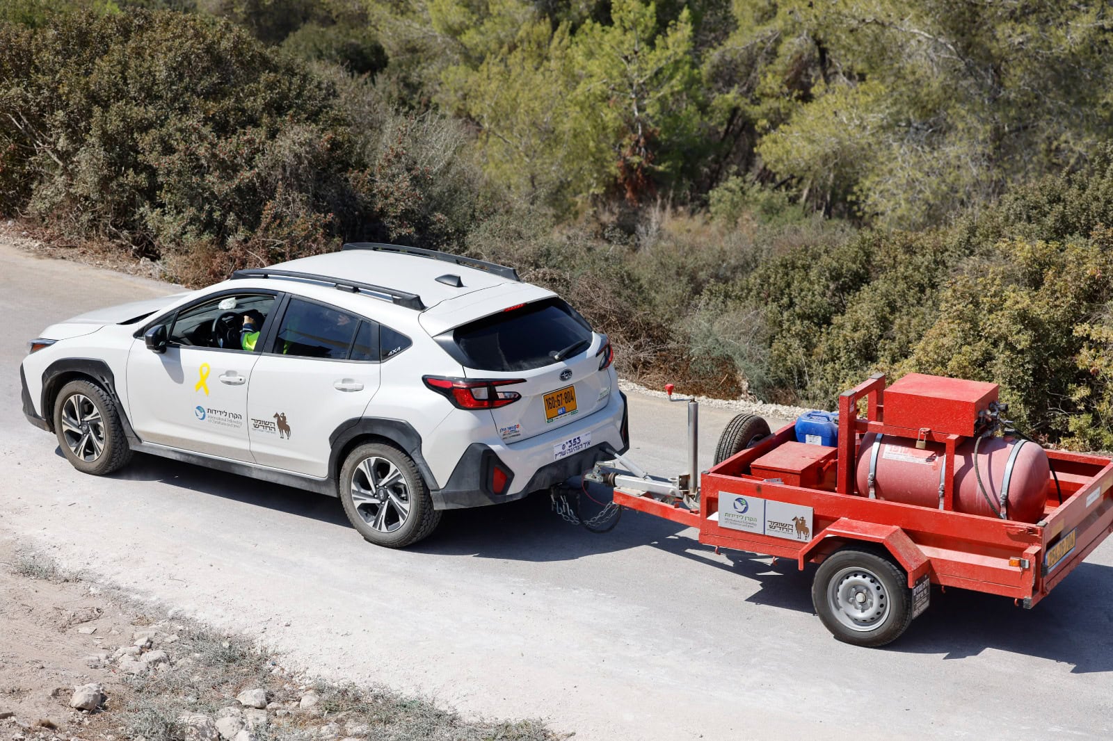 רכב שטח לבן של הקרן לידידות גורר נגרר אדום עם מכלי ציוד בדרך עפר מיוערת.